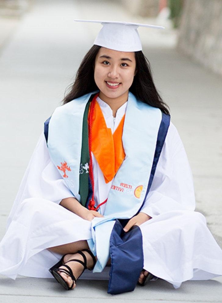 Yijie Wang wears the Pitzer white and orange graduation regalia while sitting crisscross on a walkway.